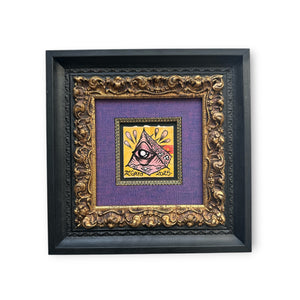 Original black and gold framed mixed media Eye Gato artwork painted on vintage film packaging, featuring the pyramid eye character in orange, black, and peach tones all within a purple mat board with gold trim.