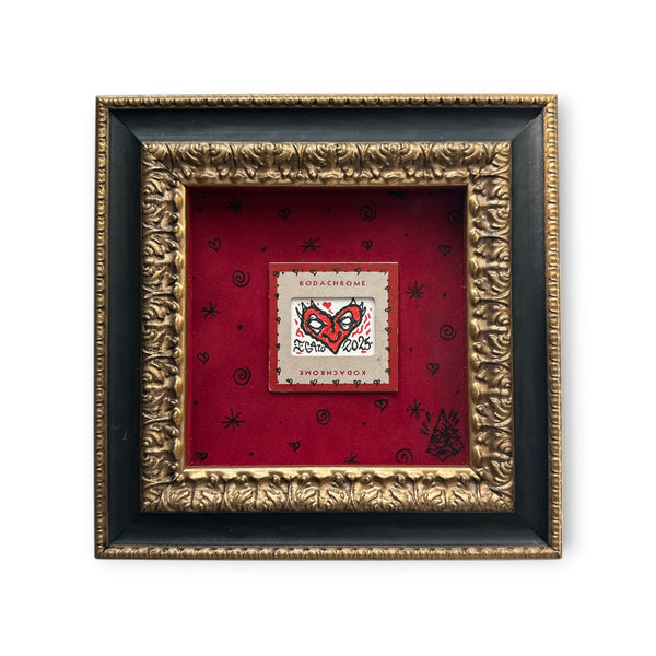 Red heart with eyes art on a piece of vintage Kodachrome film packaging. Mounted on red felt and set in a black and gold baroque-style textured wood frame.